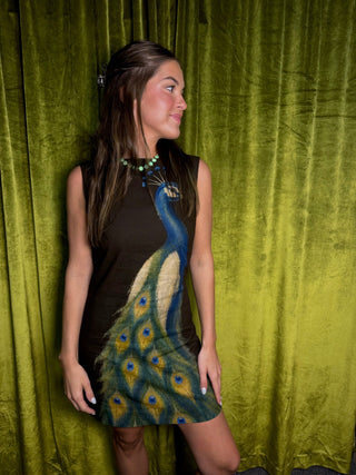 A woman wearing a sleeveless brown mini dress with a large printed peacock design on the front, standing against a textured olive green curtain backdrop.