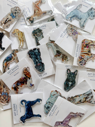 A collection of dog-shaped resin pins by Art Barke, each mounted on a white card and arranged on a light surface. The pins feature various artistic watercolor and paint-style designs in multiple colors including blue, green, brown, and purple.