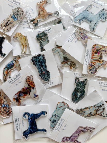 A collection of dog-shaped resin pins by Art Barke, each mounted on a white card and arranged on a light surface. The pins feature various artistic watercolor and paint-style designs in multiple colors including blue, green, brown, and purple.