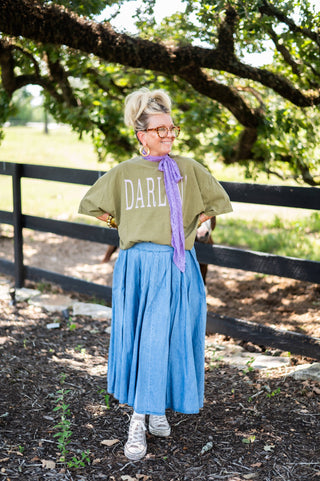 Person wearing a green shirt with 'DARLIN' printed on it, blue skirt, and white sneakers standing in a natural setting.