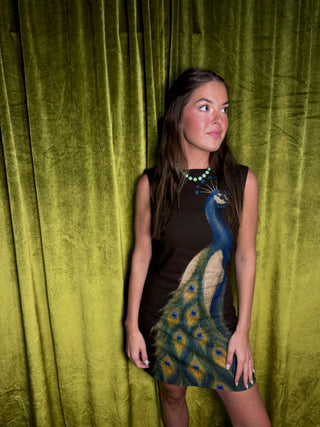 A woman posing in front of a textured olive green velvet curtain, wearing a dark brown sleeveless dress with a large peacock graphic on the front and a turquoise necklace.