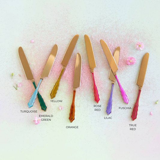 Six decorative knife-like tools with gold-toned heads and colorful handles are arranged in a row on a pale pink background with small flower petals. Each handle is labeled with a color name: turquoise, emerald green, yellow, orange, rose red, lilac, and true red.