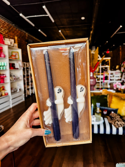 A hand holds a boxed set of two Halloween-themed candles in a retail store. The candles are dark navy blue with white ghost figures attached near the top, presented in a clear plastic window box with a kraft paper insert.