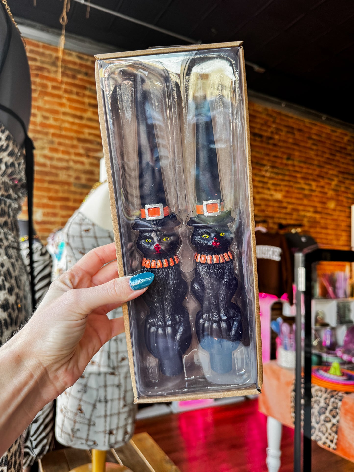 A hand with blue nail polish holds a packaged set of two black cat-shaped kitchen utensils inside a clear plastic box with a kraft paper border. The background shows a retail store interior with a brick wall and clothing mannequins.