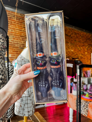 A hand with blue nail polish holds a packaged set of two black cat-shaped kitchen utensils inside a clear plastic box with a kraft paper border. The background shows a retail store interior with a brick wall and clothing mannequins.