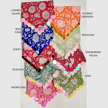 Eight patterned bandanas in different colors are arranged in a grid on a white background, each labeled with its name (Cherry Gloss, Bali Pool, Poppy, Peacock, Shortcake, Florida Oranges, Pistachio, Strawberry Fields, Lima). The bandanas feature intricate floral and traditional patterns, and many have iridescent sequin trim along the edges.