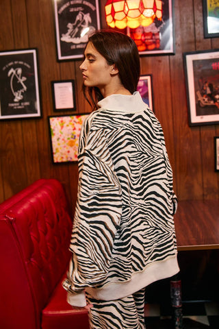 A woman wearing a black and white zebra-print sweatshirt and matching pants, standing in a retro-themed room with wood-paneled walls, framed posters, and a red tufted bench.