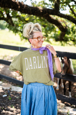 Person wearing a green 'Darlin' shirt with a blue skirt and purple scarf, standing outdoors with trees and a horse in the background.