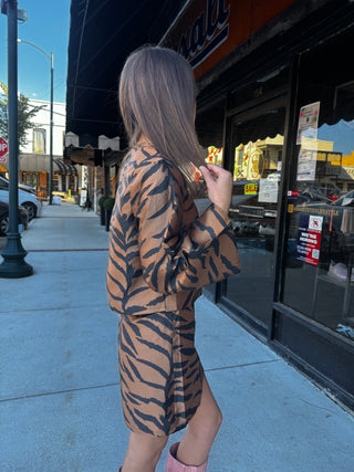 A woman stands on a city sidewalk wearing a brown and black tiger-print long-sleeve top and matching mini skirt set. She is photographed from behind, with storefront windows and a street scene in the background.