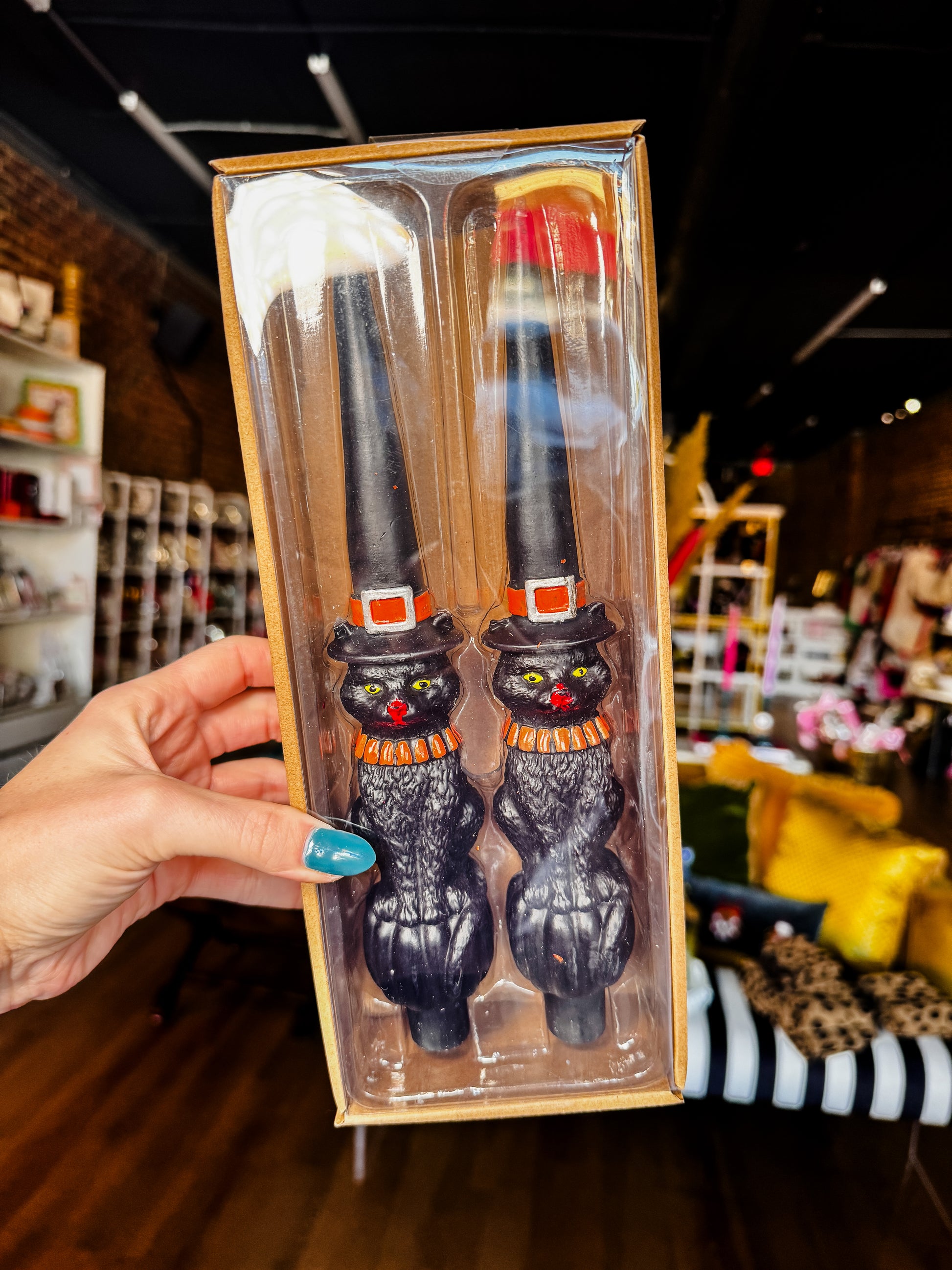 A hand with blue nail polish holds a clear plastic box containing a pair of black cat-shaped cocktail mixers with orange and yellow striped hats and scarves, set against a blurred retail store background.