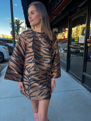 A woman wearing a brown and black tiger-print two-piece set with wide sleeves, standing on a city sidewalk in front of a storefront.