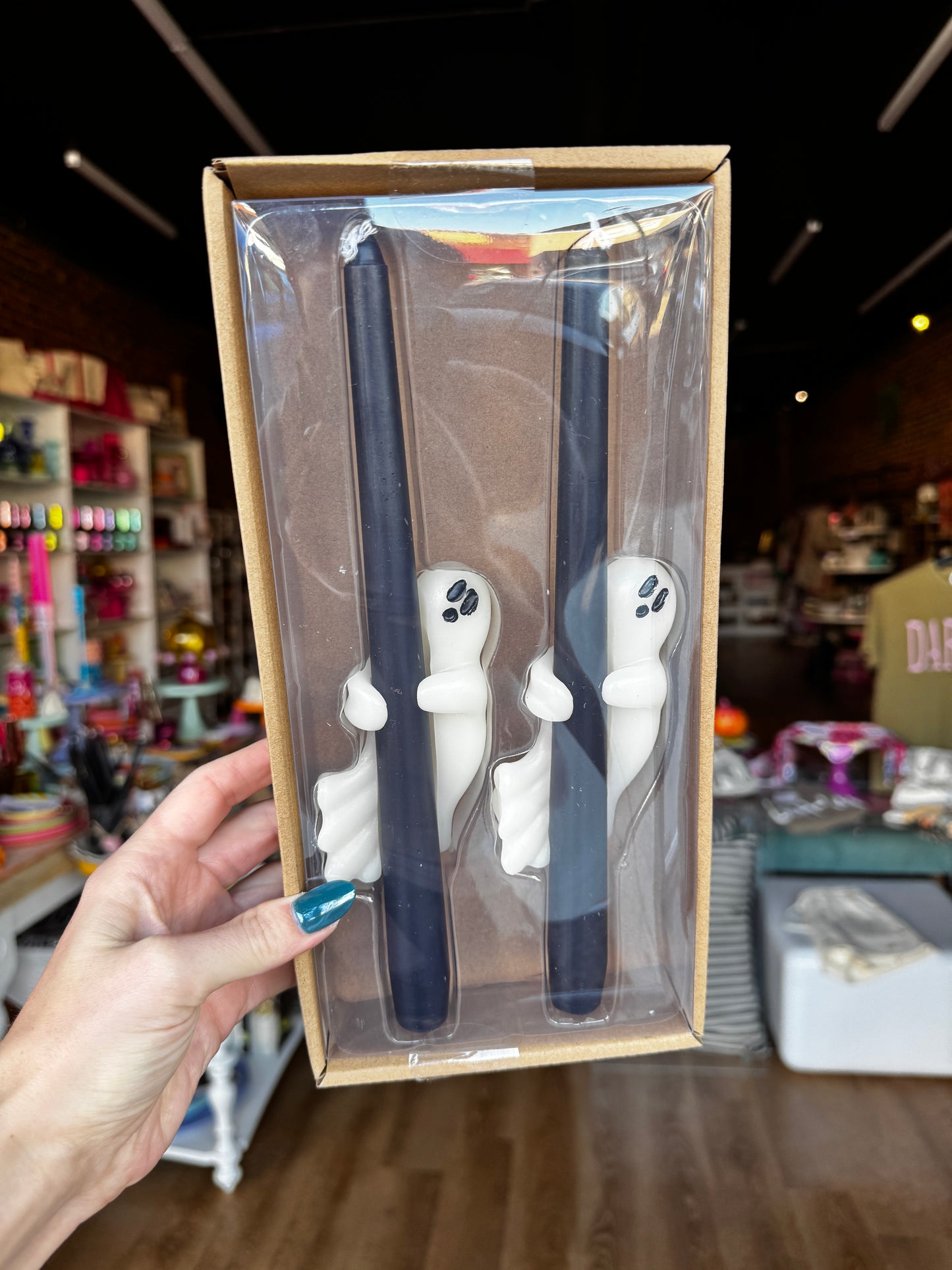A hand holds a clear plastic window box containing two dark navy blue taper candles with white ghost-shaped holders, set against a blurred retail store background.