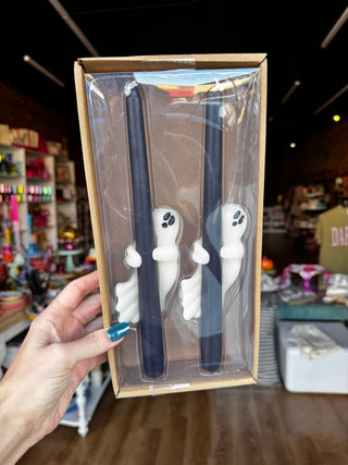A hand holds a clear plastic window box containing two dark navy blue taper candles with white ghost-shaped holders, set against a blurred retail store background.