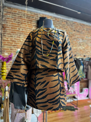 A two-piece matching set featuring a cropped long-sleeve top and a skirt, both decorated with a bold black tiger stripe pattern on a warm brown base. The set is displayed on a mannequin torso in a boutique setting with a brick wall background.