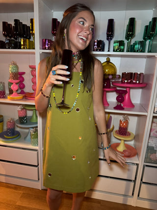 A woman wearing a sleeveless olive green dress with small colorful rhinestone embellishments, holding a dark wine glass in front of white shelves filled with colorful party supplies.