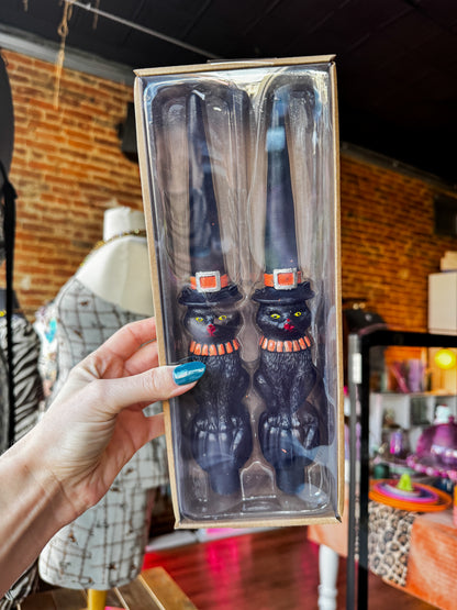 A hand holds a clear plastic box containing a pair of black cat-shaped kitchen utensils with orange and white striped accents, set against a blurred shop background.