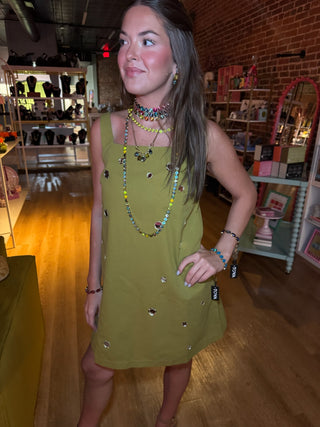 A woman modeling a sleeveless olive green dress with small metallic embellishments, standing inside a retail boutique with wooden floors and brick walls.