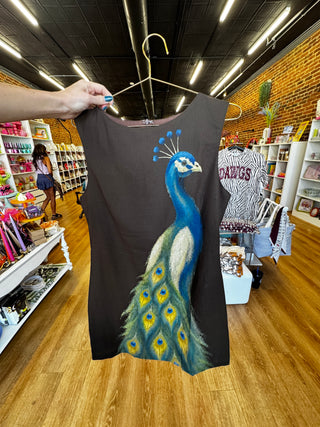 A sleeveless black tank top held up on a hanger by a person's hand inside a retail store. The shirt features a large painted peacock design in blue, green, yellow, and white on the front.