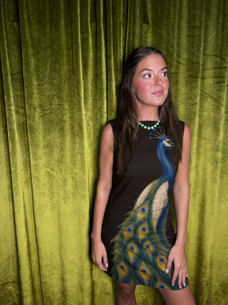 A woman posing in front of a textured olive green velvet curtain, wearing a sleeveless brown mini dress with a large printed peacock design on the front.