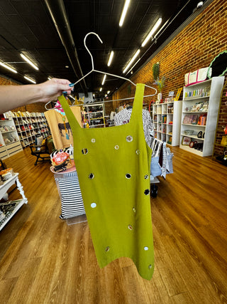 A sleeveless olive green dress with silver rhinestone embellishments hanging on a white hanger, held by a person's hand inside a clothing boutique.