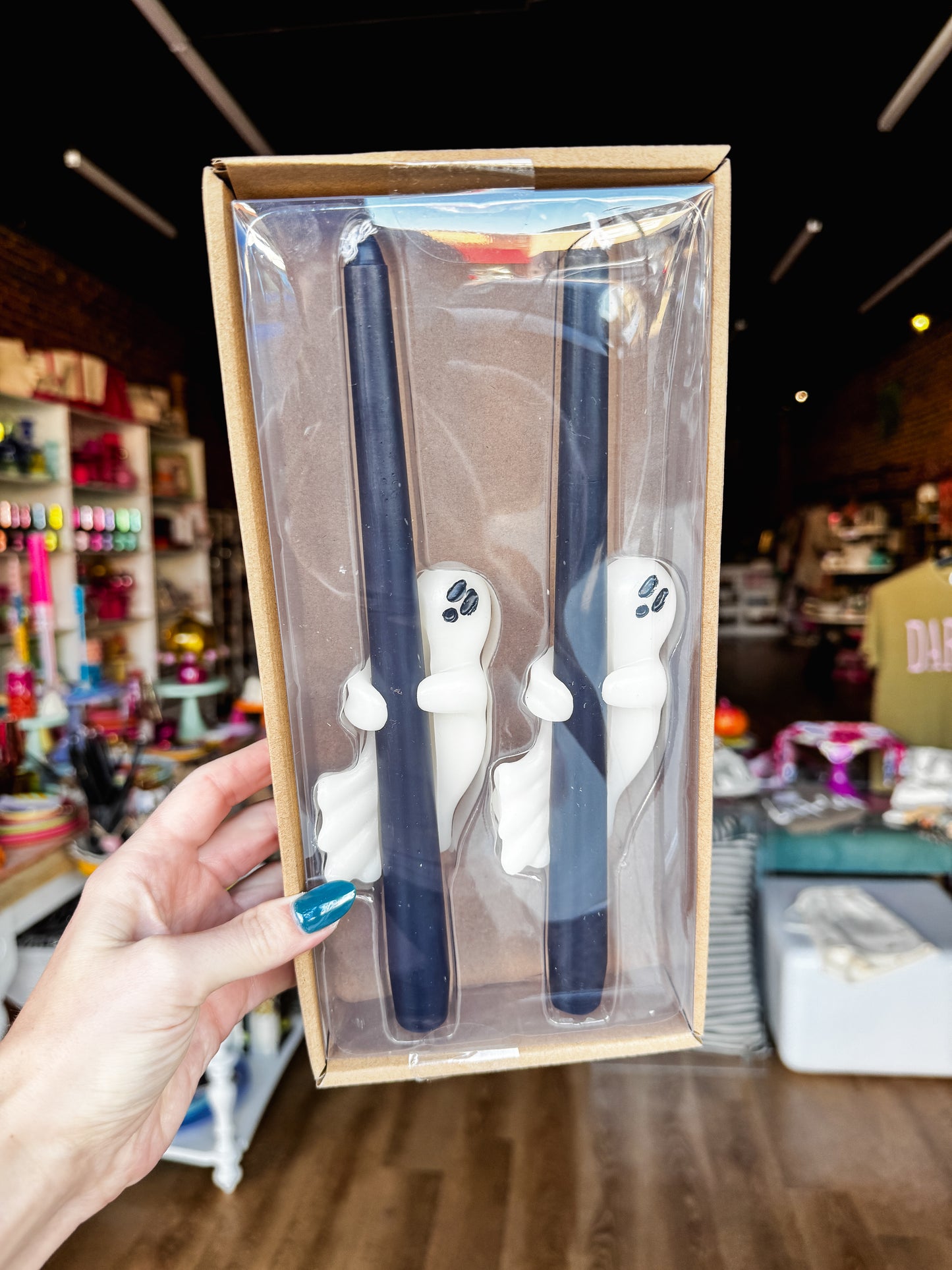 A hand holds a clear plastic gift box containing two dark blue taper candles with white ghost-shaped holders, set against a blurred retail store background.