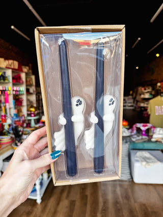 A hand holds a clear plastic gift box containing two dark blue taper candles with white ghost-shaped holders, set against a blurred retail store background.