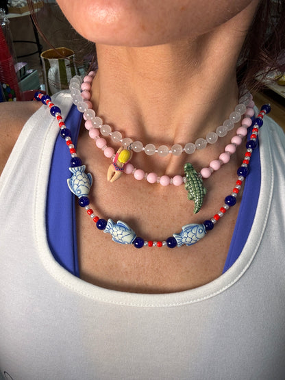 A close-up of a person's wrist wearing multiple beaded bracelets, including a blue and white fish beaded bracelet, a pink beaded bracelet, and a translucent clear beaded bracelet.