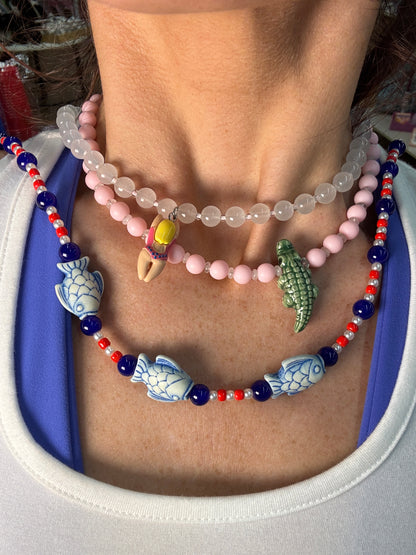 A close-up of a person's wrist wearing multiple beaded bracelets. The bracelets include a translucent white bead strand, a light pink bead strand with a small yellow and red charm, and a red, white, and blue beaded bracelet with blue fish and crocodile charms.