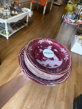 A pack of round decorative paper plates featuring a vintage-style illustration in white on a maroon background. The design includes a seated bulldog, flowers, butterflies, birds, and ornamental border motifs. Sitting on a pack of dinner plates 