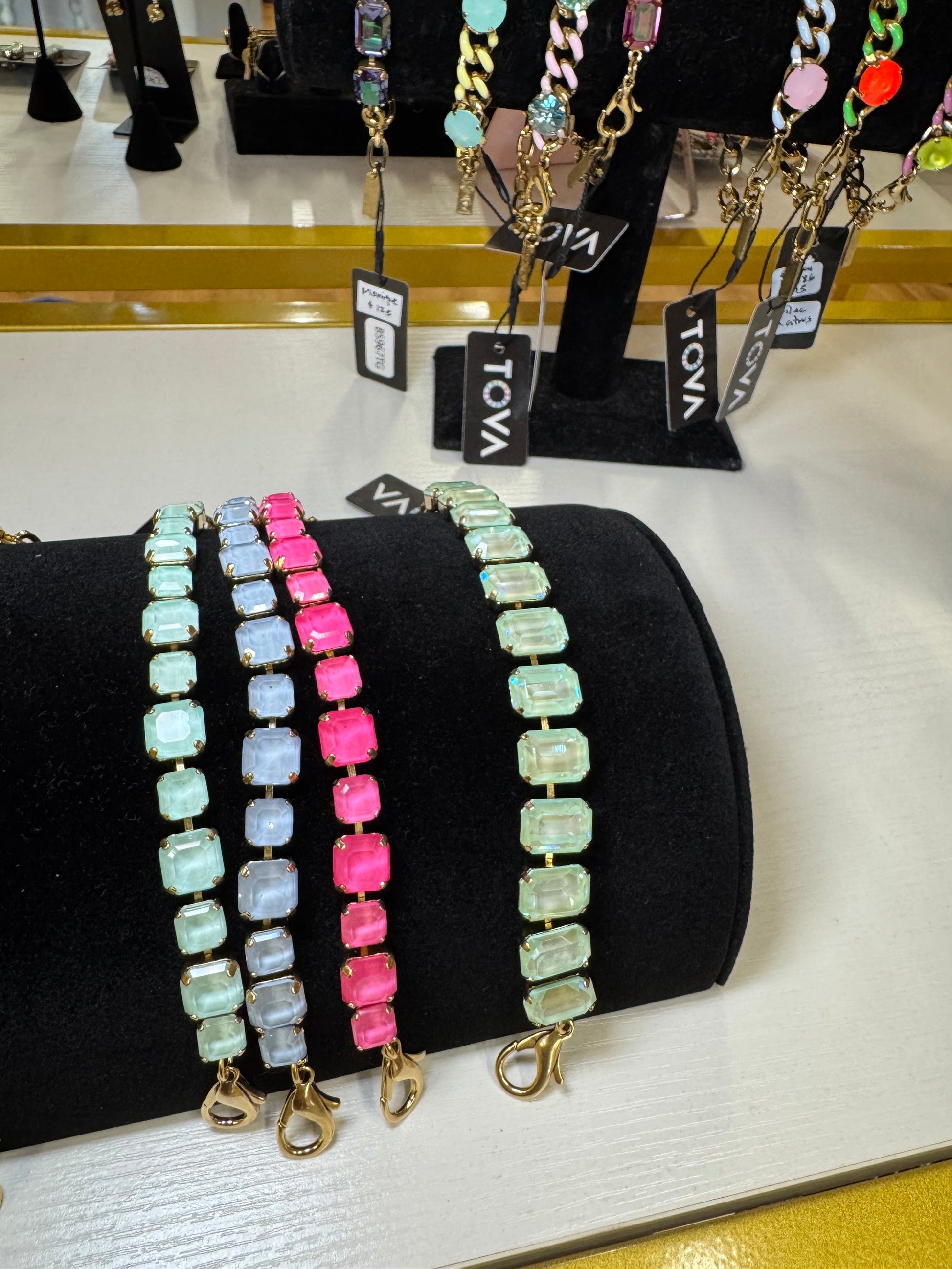 Three beaded bracelets with rectangular gemstone-like links displayed on a black velvet jewelry stand. The left bracelet is light aqua and pale blue, the middle is bright pink, and the right is mint green and pale green. TOVA
