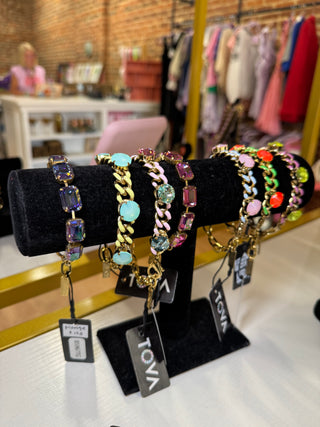 A display of colorful gemstone and chain bracelets on a black velvet stand inside a retail store. The bracelets feature gold, silver, and pink metal chains with various colored rhinestones, and have tags showing the brand 'TOVA'.