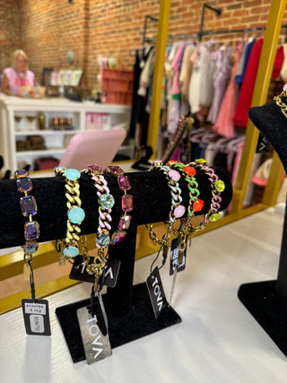 Several colorful beaded and chain-link bracelets displayed on black jewelry stands in a retail store. The bracelets feature gold-tone and silver-tone metal with multicolored rhinestones and beads. TOVA