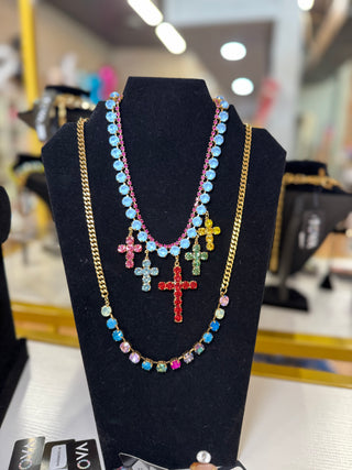 A layered necklace displayed on a black jewelry bust. The necklace features a gold chain with a long strand of light blue rhinestone beads, a shorter strand of multicolored rhinestone beads, and three dangling cross pendants in red, green, and yellow.