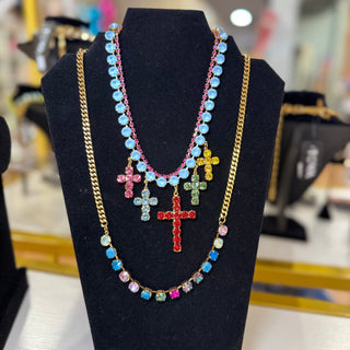 A layered necklace displayed on a black jewelry bust. The necklace features a gold chain with a long strand of light blue rhinestone beads, a shorter strand of multicolored rhinestone beads, and three dangling cross pendants in red, green, and yellow. TOVA
