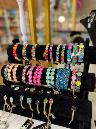 A black jewelry display stand filled with colorful beaded and rhinestone bracelets and rings, photographed in a store setting with a blurred background.