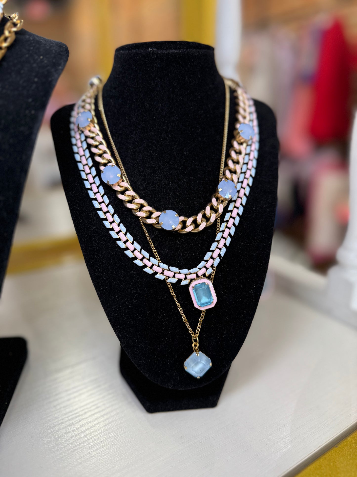 A layered necklace set displayed on a black velvet jewelry bust. The set features a gold curb chain, a white and light blue patterned chain, a gold chain with light blue oval beads, and a gold chain with a light blue faceted pendant.