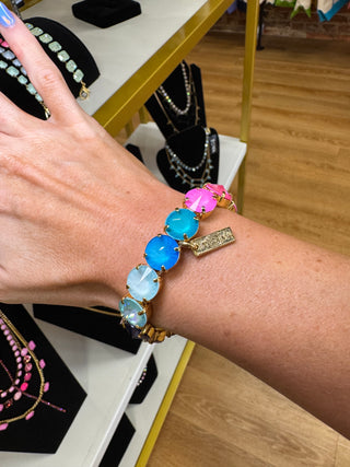 A close-up of a person's wrist wearing a gold-tone bracelet with large, round, colorful gemstone beads in shades of blue, pink, and white, alongside a small gold tag. The background shows a jewelry display shelf with other necklaces.