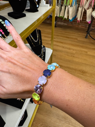 A gold-tone bracelet with seven colorful oval gemstones in shades of green, purple, blue, and pink, worn on a person's wrist in a jewelry store.