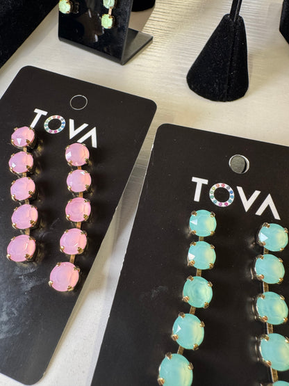 Two pairs of Tova brand statement earrings displayed on black cards. The left pair features round pink rhinestones, and the right pair features oval light blue-green rhinestones, both with gold-tone settings.