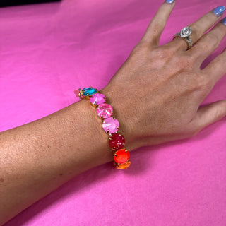 A colorful beaded stretch bracelet with pink, red, orange, and turquoise beads on a gold-tone band, shown worn on a person's wrist against a bright pink background.