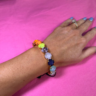 A multicolor beaded bracelet with oval gemstones in orange, yellow, green, purple, white, and blue, worn on a person's wrist against a bright pink background.