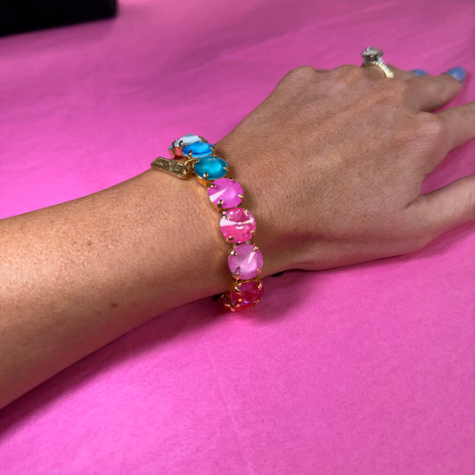 A close-up of a person's hand wearing a gold-toned beaded bracelet with alternating pink and turquoise blue oval beads, resting on a bright pink surface.