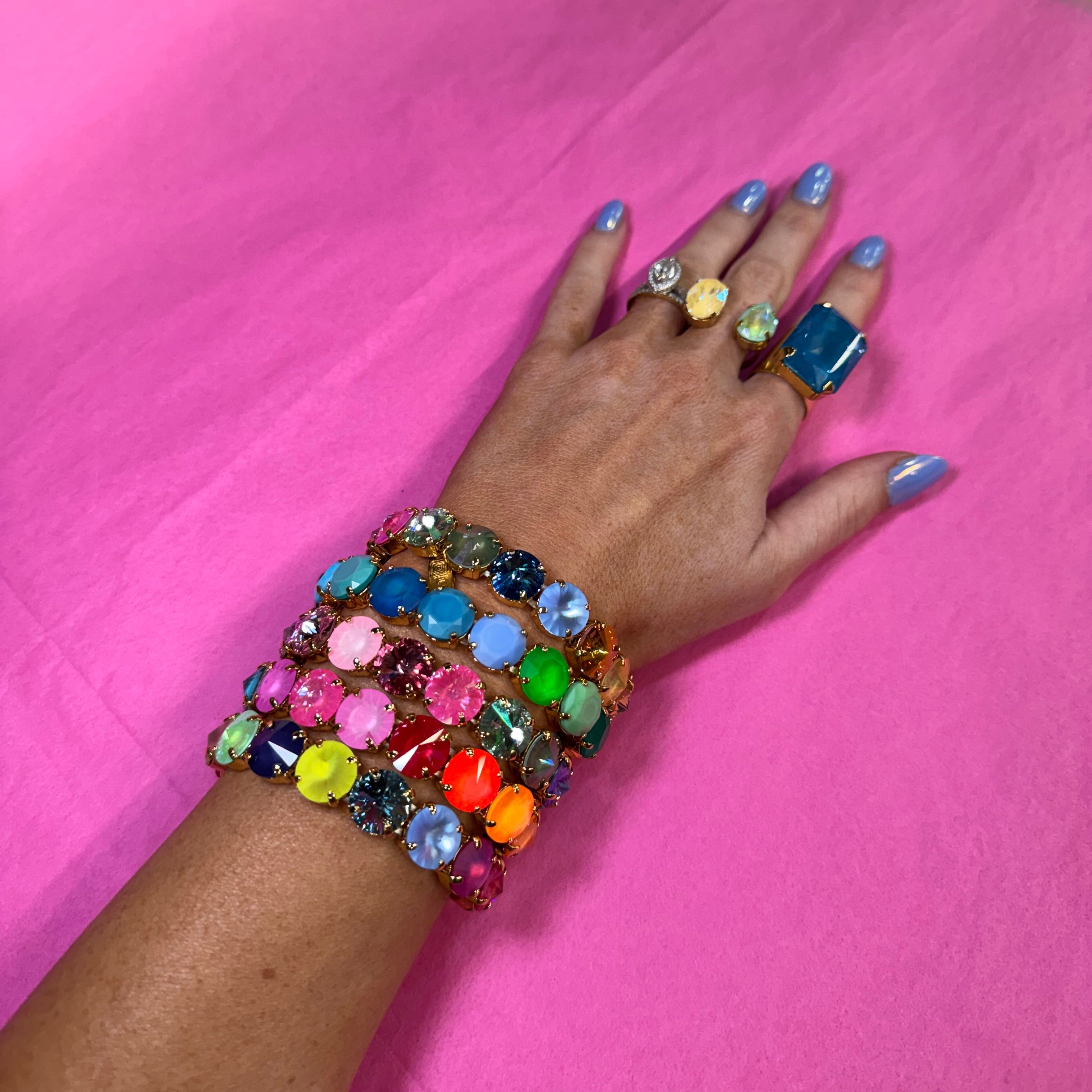 A close-up of a person's hand and wrist wearing multiple colorful gemstone bracelets and rings, resting on a bright pink background.