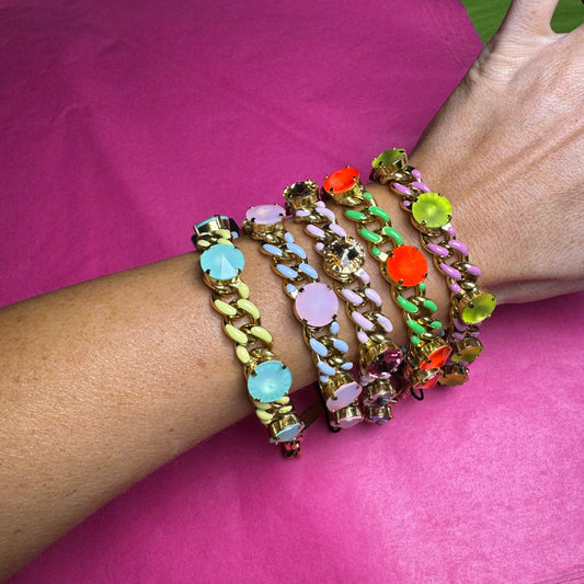 A set of five chunky gold-tone chain bracelets with colorful faux gemstones, worn stacked on a person's wrist against a bright pink background.