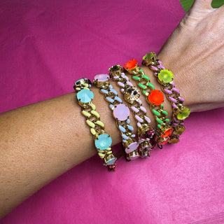A set of five chunky gold-tone chain bracelets with colorful faux gemstones, worn stacked on a person's wrist against a bright pink background.