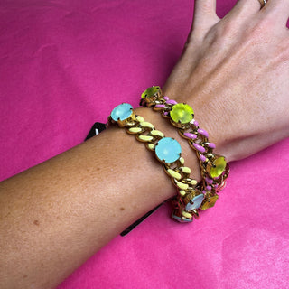A close-up of a person's wrist and hand wearing three colorful chain bracelets with round light blue and lime green stones, set against a bright pink background.