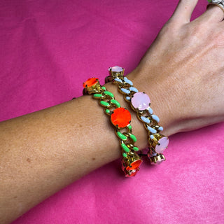 Three colorful chain link bracelets with gemstone accents worn on a person's wrist, set against a bright pink background. TOVA