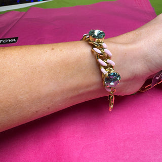 A gold chain bracelet with pink enamel links and green crystal accents, worn on a person's wrist over a bright pink background. TOVA