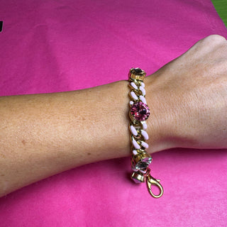 A gold-tone chain bracelet with white pearl-like links, a pink faceted crystal centerpiece, and a dangling pink crystal charm, shown worn on a person's wrist against a bright pink background. TOVA