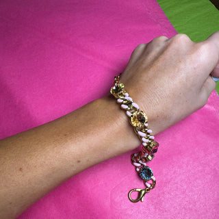 A close-up of a person's wrist wearing a colorful chain bracelet with gold-tone links, white beads, and small multicolored stones, set against a bright pink and green background. TOVA